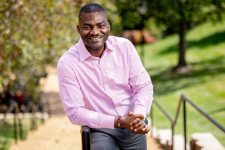 Olu Owoeye, Ph.D., leans against a railing for a photo on a bright sunny day. He is smiling and wearing a long-sleeve pink shirt.